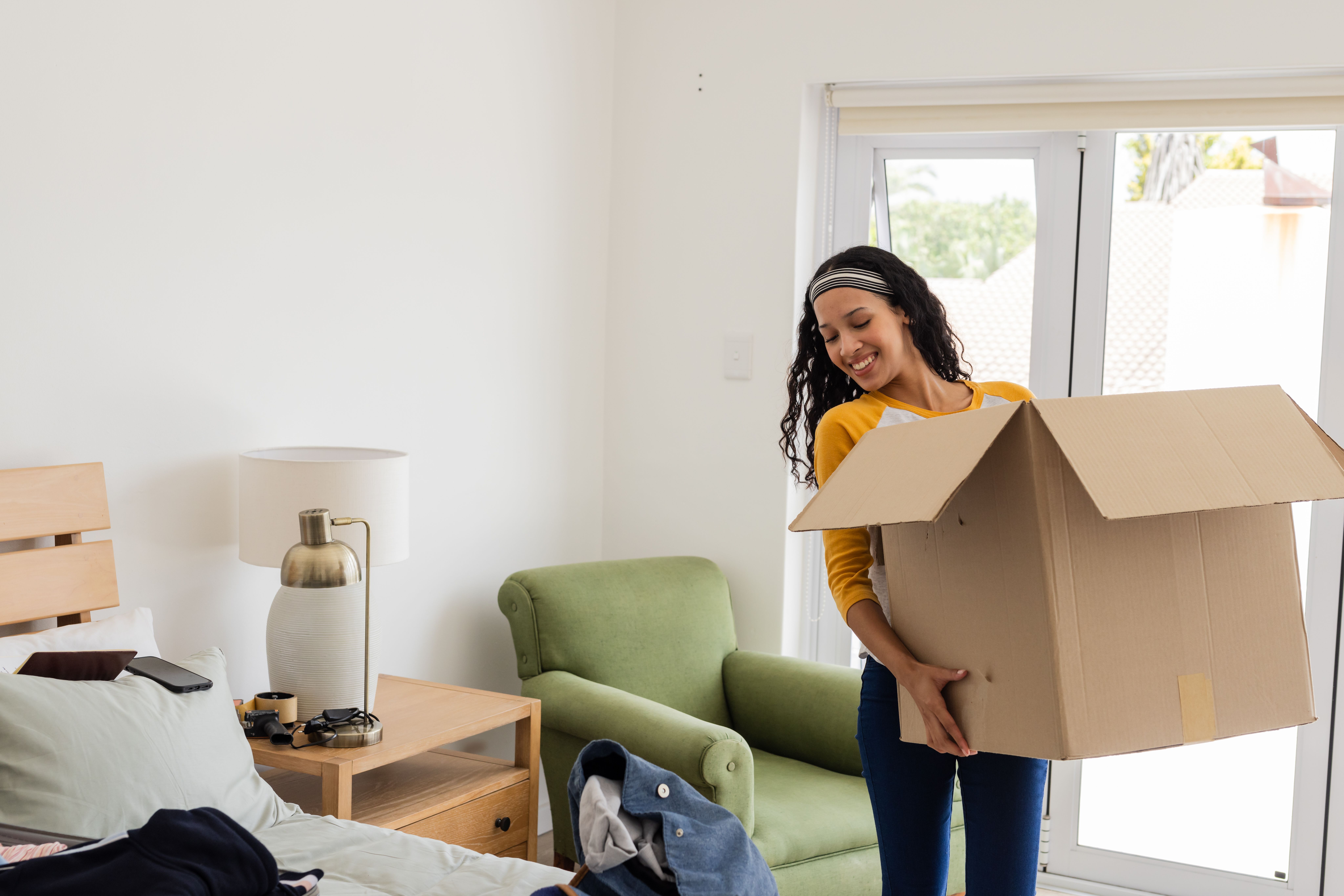 Unpacking cardboard box in bedroom, African American woman organizing belongings, smiling